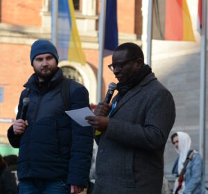Karamba hält eine Rede auf dem Marktplatz von Halle; er hat ein Mikrofon und einen Sprechzettel in der Hand. Neben ihm steht ein junger Mann, der dolmetscht.